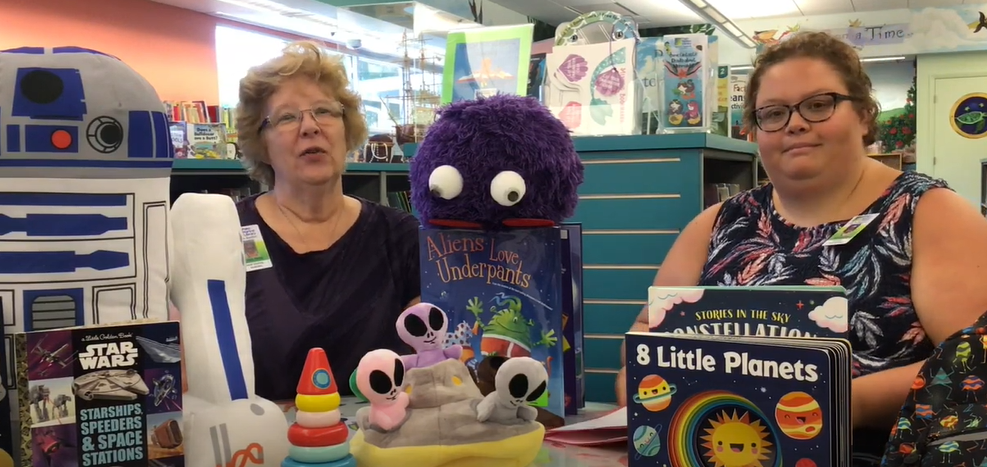 Two librarians sit with books, C3PO and alien dolls. 