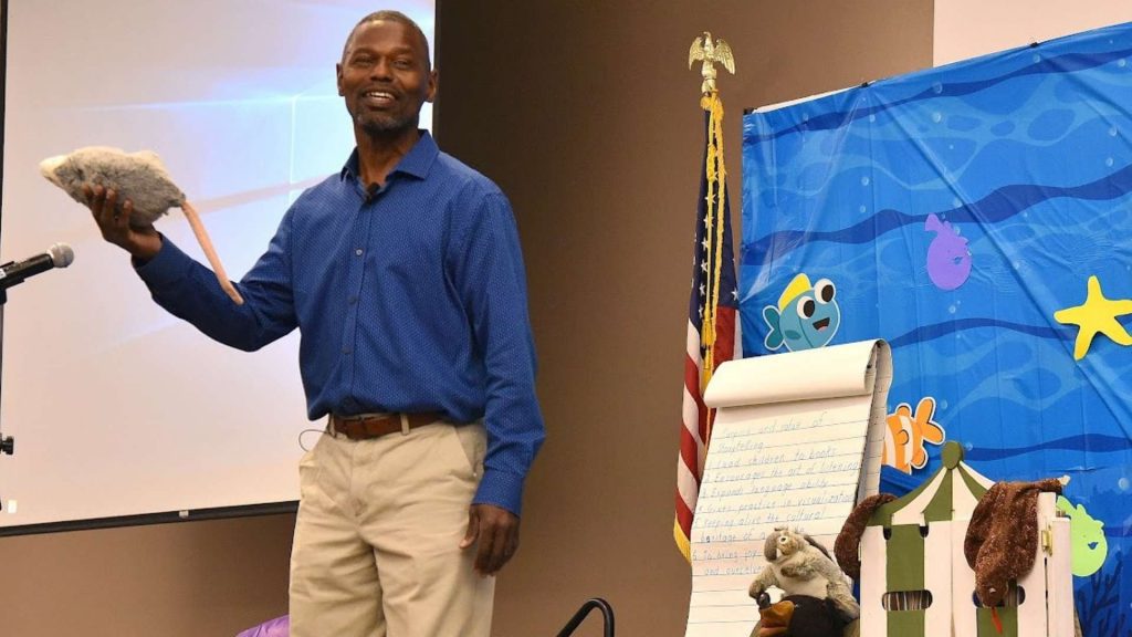 Storyteller stands in a stage with a kid-friendly background and stuffed animals