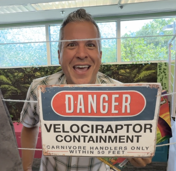 A man smiles behind a Danger: Velociraptor Contaiment sing.
